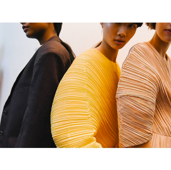 Three models photographed in profile wearing Jil Sander pieces featuring sculptural, accordion-pleated sleeves in mustard yellow and tan tones alongside a dark tailored jacket.