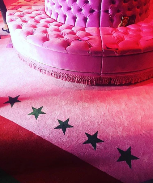 A photograph of a round, tufted pink velvet sofa with fringed trim positioned on a plush carpet featuring black stars in gradations of pink and red.